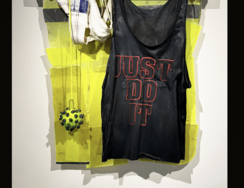 A mixed media artwork by Tom Hill titled "Finish Line" from 2024, featuring a black tank top with "Just Do It" text, mounted with various materials like PVC tubing, lumber, and athletic gear.